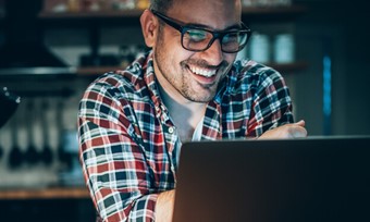 A man with glasses smiles while using his laptop, reflecting a positive and focused work environment.